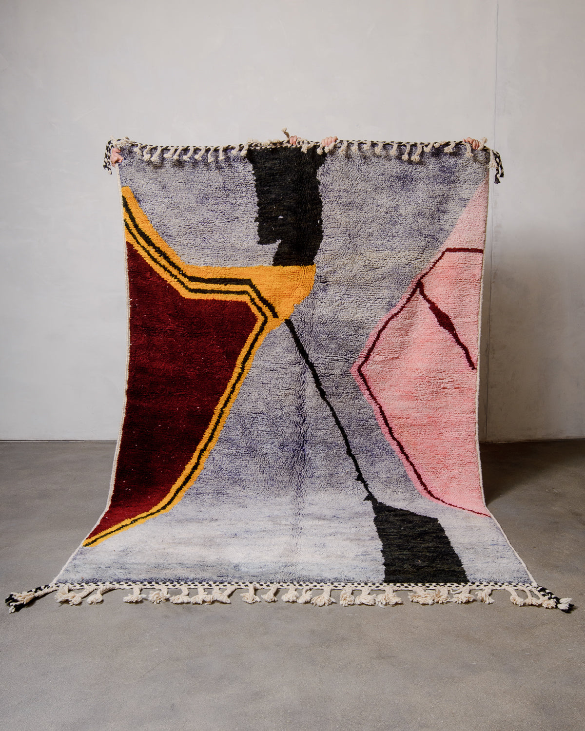 Berberlin special edition berber rug featuring lavender, pink and burgundy being held up in front of white wall and concrete floor.
